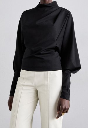 Haut noir à manches longues avec col montant et manches bouffantes, associé à un pantalon taille haute blanc cassé avec des coutures verticales.