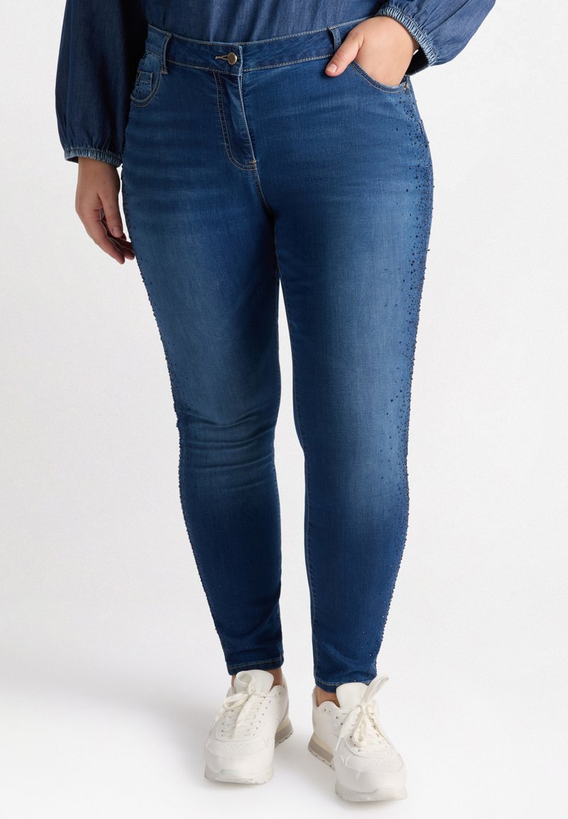 Jeans ajustados de mezclilla azul con textura acanalada, que cuentan con tachuelas decorativas a lo largo de las costuras laterales. Combinados con zapatillas blancas.