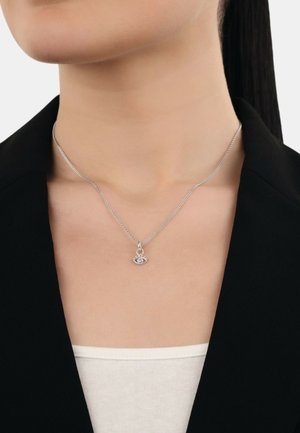 Collana d'argento con una catena delicata e un pendente a forma di occhio con una pietra centrale, indossata sopra una blusa chiara e sotto un blazer nero.