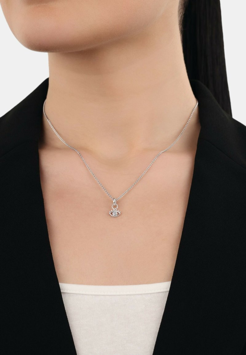 Collana d'argento con una catena delicata e un pendente a forma di occhio con una pietra centrale, indossata sopra una blusa chiara e sotto un blazer nero.
