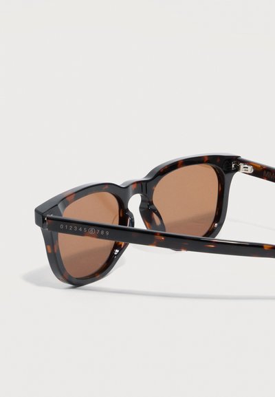 Γυαλιά ηλίου με σχέδιο χελώνας, καφέ φακούς, στρογγυλό πλαίσιο και αριθμητική χάραξη στο πλαϊνό μπράτσο, με φως φόντο.