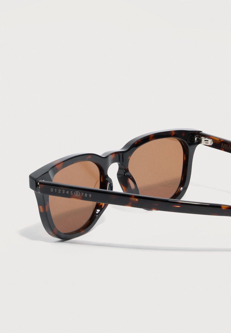 Occhiali da sole tartarugati con lenti marroni, montatura rotonda e incisione numerica sull'asta contro uno sfondo chiaro.