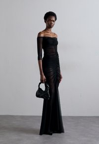 Rochie neagră cu umerii la vedere, cu detalii plisate, țesătură transparentă și despicătură. Modelul ține o poșetă mică neagră cu un design curbat.