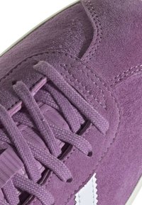 Violet zamšādas kedas ar strukturētu virsmu, aprīkotas ar plakaniem šņorēm un baltām logo akcentiem. Ieskaitot izšūtu detaļu gar šuvēm.
