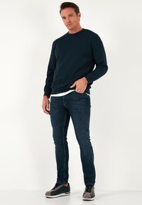 Tengeri kék pulóver kerek nyakkal, sötét farmernadrággal párosítva. A modellen szürke és tengeri kék sportcipő van, amelyek minimalista dizájnnal büszkélkednek.
