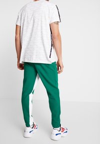 Man i gröna och vita träningsoverallsbyxor, vit tryckt kortärmad skjorta och vita sneakers med röda och blå detaljer, sedd bakifrån.