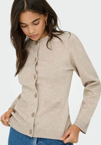 Cardigan en tricot beige avec une fermeture à boutons, tissu texturé, poignets et ourlet côtelés, et deux poches avant. Le modèle tient l'ourlet tout en regardant vers le bas.