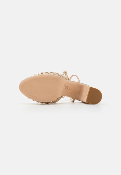 Sandalía de tacón beige con suela lisa, diseño de tiras y cinta delgada en el tobillo con un pequeño broche. Plantilla texturizada para mayor agarre.