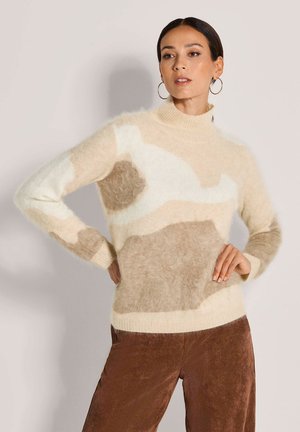 Femme aux cheveux foncés portant de grandes boucles d'oreilles en anneau, un pull beige et blanc en tissus doux, et un pantalon en velours marron, posant avec une main sur la hanche.