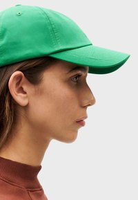 Groene honkbalpet van stof, voorzien van een gebogen klep en gestikt detail, weergegeven vanaf de zijkant naast een effen bovenstuk.