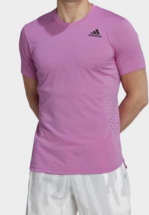 Muž nosící přiléhavé fialové tričko Adidas s ventilačními otvory po stranách a bílé šortky s vzorem.