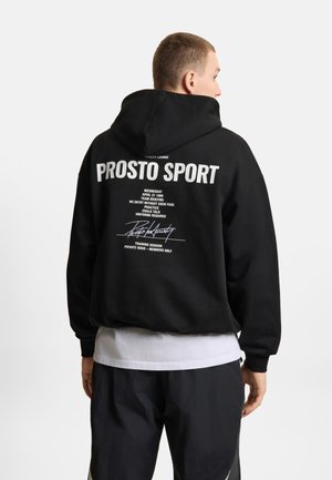 Mężczyzna noszący czarną bluzę z kapturem z białym napisem "PROSTO SPORT" oraz mniejszym białym napisem na plecach, połączoną z czarnymi spodniami i białą koszulą.