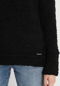 Pull noir texturé avec un design lâche et duveteux. Présente un petit logo en argent près de l'ourlet. Associé à un jean en denim.