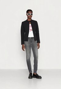 Black quilted jacket, graphic white t-shirt, gray slim-fit jeans, and chunky black shoes. White sunglasses complete the look. Simple background.