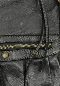 Détail d'un sac en cuir noir avec une fermeture éclair dorée visible, deux boutons-pression en laiton et une surface texturée avec des coutures contrastantes.