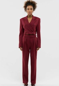 Vrouw met krullend haar in een bordeauxrode dubbelbreasted cropped blazer en bijpassende hoog getailleerde broek, staand tegen een witte achtergrond.