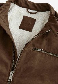 Brūna zamšādas jaka ar baltu vilnas oderi, priekšējais rāvējslēdzis daļēji atvērts, un redzama rāvējslēdzēja krūšu kabata.