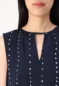 Marinebluse mit weißen Punkten, mit einem Schlüsselaugen-Ausschnitt, kurzen Ärmeln und einer weichen, leichten Stofftextur.