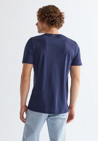Mand set bagfra iført en ensfarvet mørkeblå kortærmet t-shirt og lyseblå jeans mod en hvid baggrund.