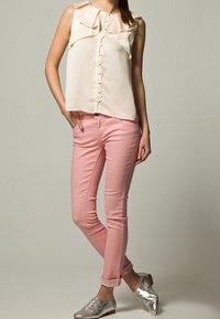 Hellbeige ärmellose Bluse mit einem rüschendekolleté, durchgehender Knopfleiste; kombiniert mit pinken Slim-Fit-Jeans und silbernen Schuhen. Einfaches Design.