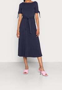 Robe midi bleu marine à manches courtes, détails noués à la taille et texture lisse, associée à des mules à talons hauts roses.