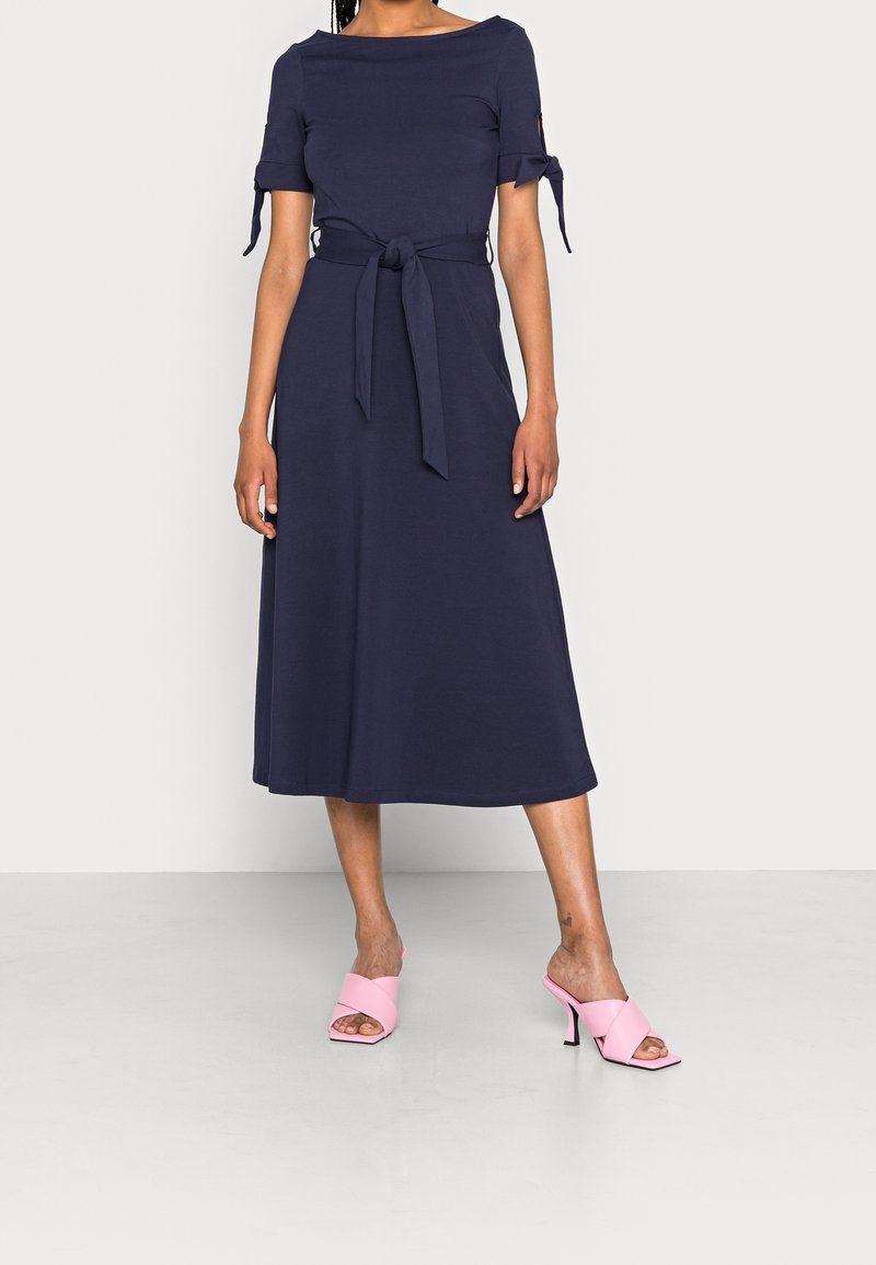 Robe midi bleu marine à manches courtes, détails noués à la taille et texture lisse, associée à des mules à talons hauts roses.