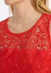 Rotes Spitzenoberteil mit aufwendigen Blumenmustern, gewelltem Ausschnitt und einer mehrschichtigen, durchsichtigen Gestaltung mit glatter Unterlage.