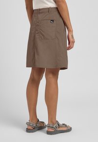 Jupe en coton marron, arrivant au genou, avec une poche latérale portant une étiquette, associée à des sandales à motif. Texture lisse et design simple.