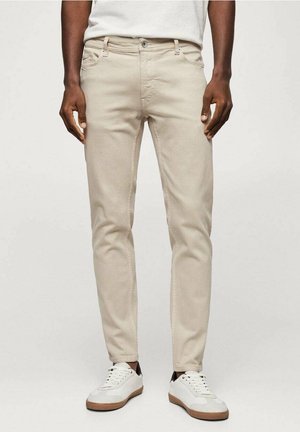 Mann trägt beige, schmal geschnittene Hose, weiße Sneakers mit Gummisohlen und ein weißes T-Shirt, steht vor einem schlichten Hintergrund.