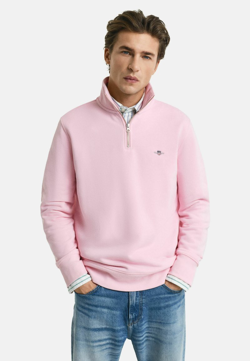 Felpa rosa chiaro con colletto a zip, caratterizzata da polsini e orlo a coste. Indossata sopra una camicia a righe, con un piccolo logo sul petto.