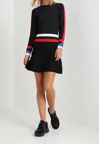 Nainen, jolla on päällään musta hame ja pitkähihaiset mustat paidat, joissa on punaisia, valkoisia ja sinisiä raitoja, kävelee mustissa lenkkareissa valkoisella lattialla.