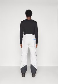 Pantalones de esquí blancos con detalles grises, cinturilla elástica y cremalleras en los lados. Combinados con una camiseta negra de manga larga, vistos desde la parte posterior.
