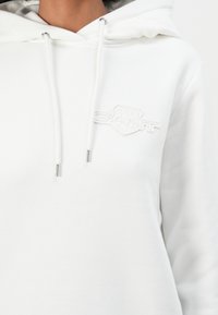Sweat à capuche blanc avec cordons et logo brodé subtil sur la poitrine gauche, porté par une personne avec le cou et les épaules visibles.