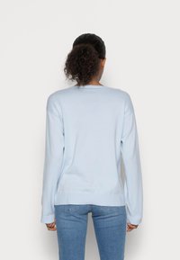 Frau mit lockigem, nach hinten gebundenem Haar, die einen hellblauen Langarmpullover und blue Jeans trägt, mit dem Rücken zu einer schlichten Hintergrund.