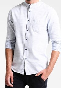 Chemise bleu clair à boutons avec col mao, texture à motifs, une poche poitrine unique et boutons bleu foncé. Manches longues retroussées.