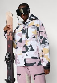 Valge suusajakk roosa, halli ja kuldse abstraktse kamuflaažimustriga, millel on lukuga taskud ja kapuuts. Saatjaks mustad suusad.