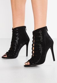 Zwarte enkellaarsjes met open tenen, met een fonkelende textuur, vetersluiting aan de voorkant en hoge stilettohakken. Ritssluiting aan de achterkant.