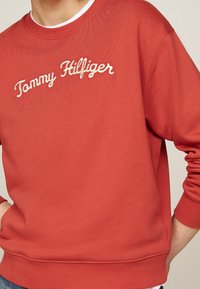 Roter Sweatshirt aus Baumwolle, mit Rundhalsausschnitt und gerippten Bündchen, auf dessen Vorderseite "Tommy Hilfiger" in weißer Schreibschrift gestickt ist.