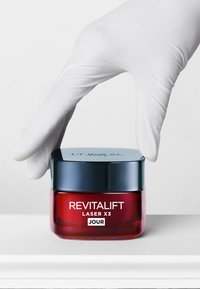 Un pot rouge et transparent avec un couvercle bleu foncé étiqueté "REVITALIFT LASER X3 JOUR". Une main gantée tend la main vers le couvercle. Surface lisse.