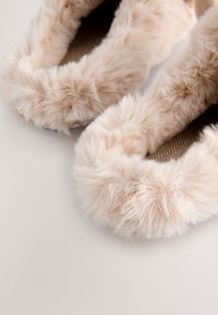 Chaussons en fausse fourrure beige avec un bord moelleux et duveteux et une semelle intérieure texturée tissée. La semelle est lisse, offrant une apparence douce et un confort.