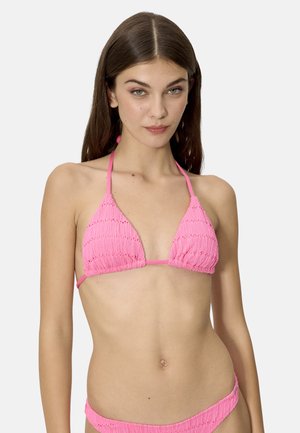 Joven con cabello largo y castaño que lleva un bikini rosa texturizado de triángulo y braguitas a juego sobre un fondo liso.