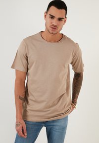 Tricou bej cu mânecă scurtă, cu guler rotund, textură moale și design fără cusături. Purtați cu blugi albaștri, evidențiind detalii ale tatuajului de pe braț.