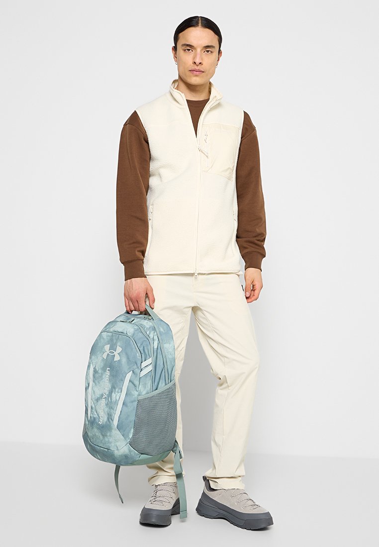 Gilet in pile color crema, camicia marrone a maniche lunghe, pantaloni chiari e scarpe grigie. Zaino azzurro chiaro con tasca laterale in rete e logo.