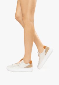 Le sneakers presentano una base bianca con accenti color sabbia e un tallone dorato texturizzato. I lacci sono di un rosa chiaro, evidenziando il design minimalista.