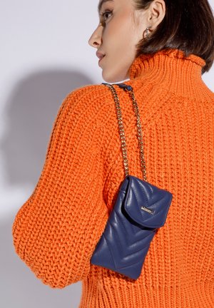 Mujer con cabello corto y oscuro que lleva un suéter grueso de punto naranja y un pequeño bolso acolchado azul marino con una correa de cadena dorada.