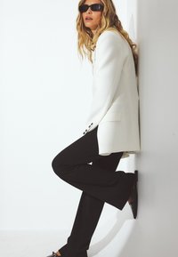 Bela blazer z črnimi gumbi, v kombinaciji s krojenimi črnimi hlačami in konicami črnih balerink. Obleka ima brezšiven, oster dizajn.