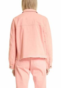 Kvinde iført en lys pink denimjakke og matchende bukser, vist fra siden med lige, blond hår.