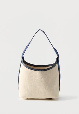 Μικρή μπεζ υφασμάτινη τσάντα με μπλε δερμάτινη επένδυση και έναν μπλε ιμάντα, όρθια σε επίπεδη επιφάνεια με απλό φόντο.