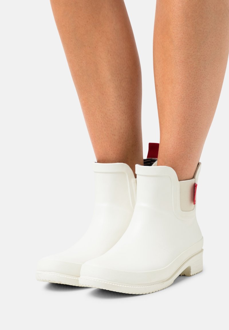 Bas des jambes portant des bottines de pluie blanches avec des languettes rouges sur un fond clair uni.