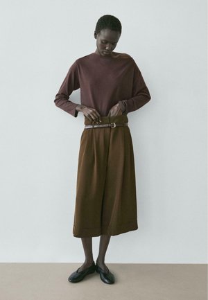 Donna con indosso un top a maniche lunghe color mogano e pantaloni culotte marroni a gamba larga, che si sistema una cintura, in piedi davanti a uno sfondo chiaro e semplice.